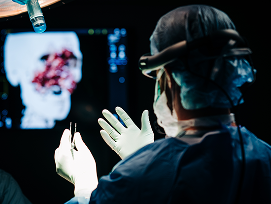 The back of a man in medical gown and scrub cap with VR headset holding gloved hands out in front of him with screen in the background with a skull x-ray
